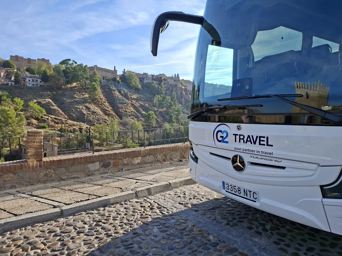Autocar blanco con rotulación G2 Travel, visto de frente junto a un paisaje monumental.