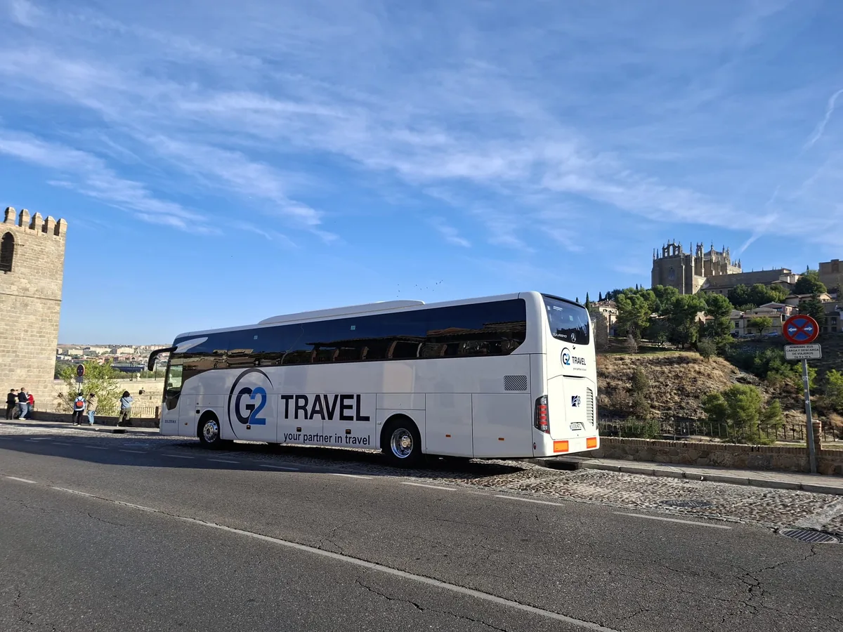 Vista lateral de un autocar blanco con rotulación G2 Travel junto a un recinto histórico.