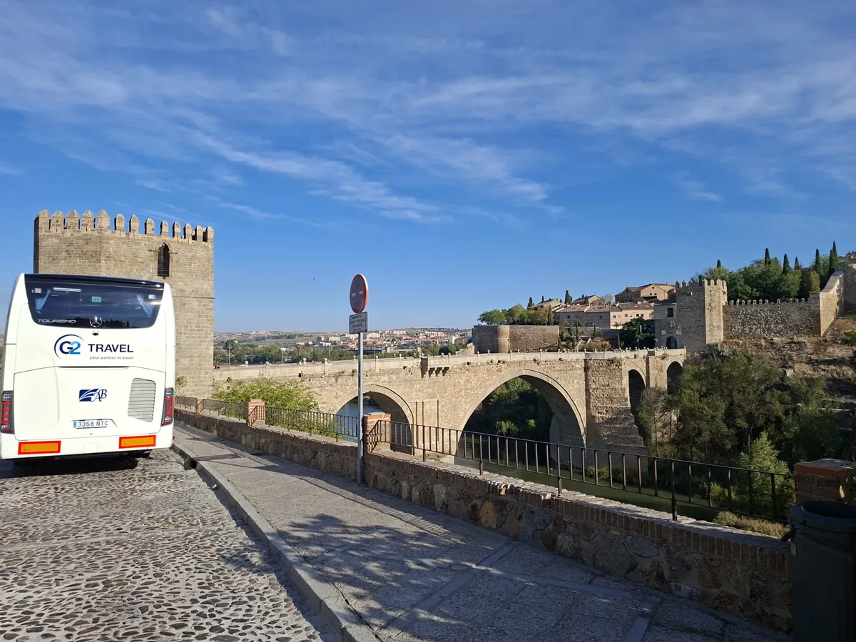 Vista trasera de un autocar blanco con rotulación G2 Travel junto a un puente de piedra.