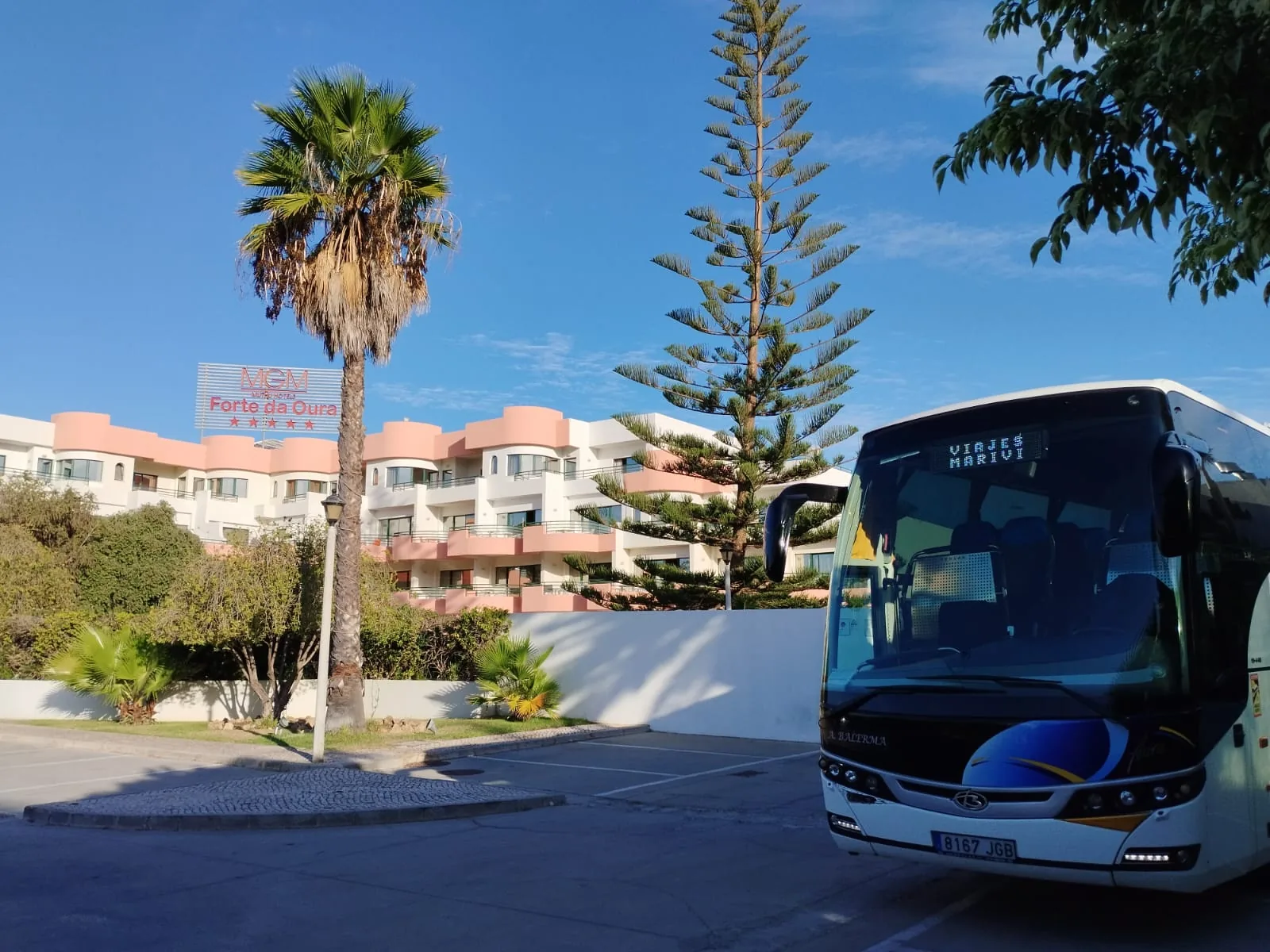 Vista frontal de un autocar Balerma con decoración azul y amarilla junto a un hotel con palmeras.
