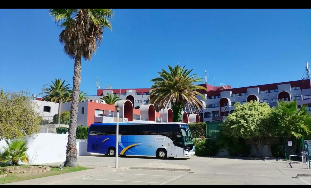 Vista general de un autocar Balerma con decoración azul y amarilla en un aparcamiento junto a un hotel.